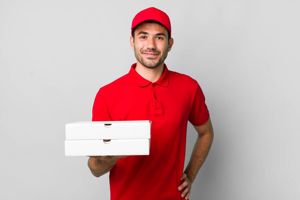 Genç, İspanyol bir adam mutlu bir şekilde gülümsüyor, eli kalçasında ve kendinden emin. Pizza teslimatı kavramı - Fotoğraf, Görsel