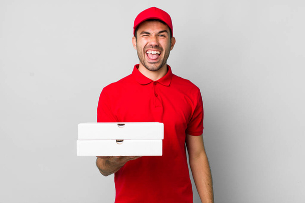 young adult hispanic man with cheerful and rebellious attitude, joking and sticking tongue out. pizza delivery concept - Фото, изображение
