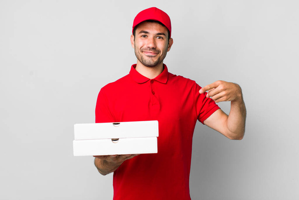 young adult hispanic man smiling cheerfully, feeling happy and pointing to the side. pizza delivery concept - Foto, afbeelding