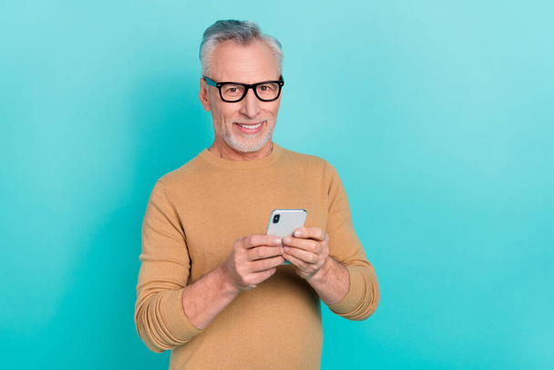 Portrait of cheerful aged man hold telephone typing texting post comment isolated on teal color background - Fotó, kép