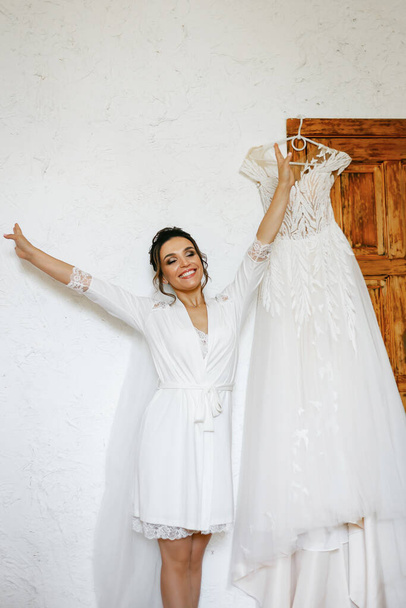 Una novia con una túnica blanca posando cerca de su vestido de novia. - Foto, Imagen