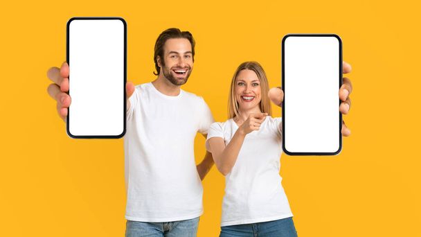 Glückliche Millennial-Europäer in weißen T-Shirts zeigen mit dem Finger auf Smartphones mit leerem Bildschirm - Foto, Bild