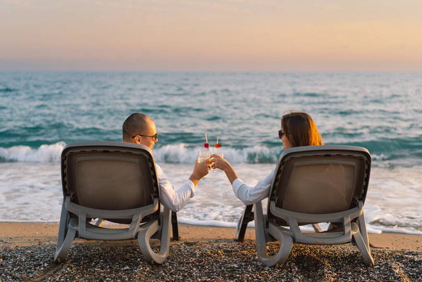 Para siedzi w salonie krzesła na letnie wakacje cieszy się zachodem słońca w pobliżu morza z szklanym koktajlem - Zdjęcie, obraz