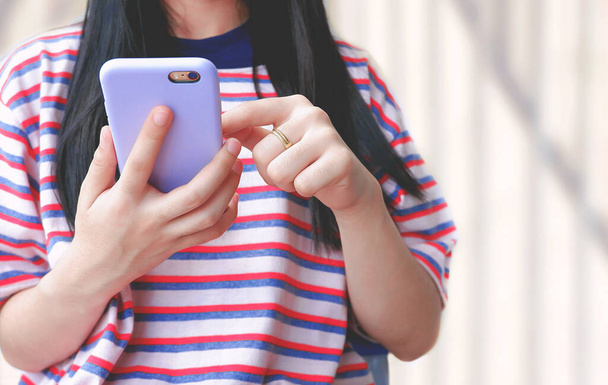 Close up of female teenager in striped t-shirt using smartphone on blurred white wall background in outdoor area - Zdjęcie, obraz