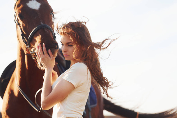 Beautiful sunshine. Young woman standing with her horse in agriculture field at daytime. - Foto, afbeelding