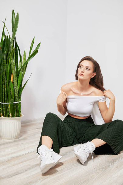 young beautiful girl in green pants near a green plant sitting on the floor - Foto, imagen