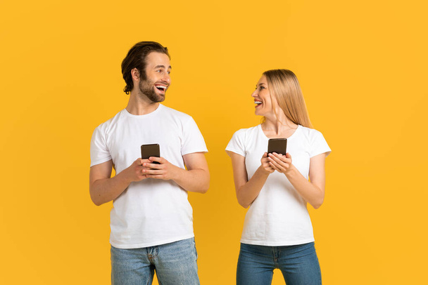 Веселые кавказские женщины и мужчины в белых футболках смотрят друг на друга, держат телефоны - Фото, изображение