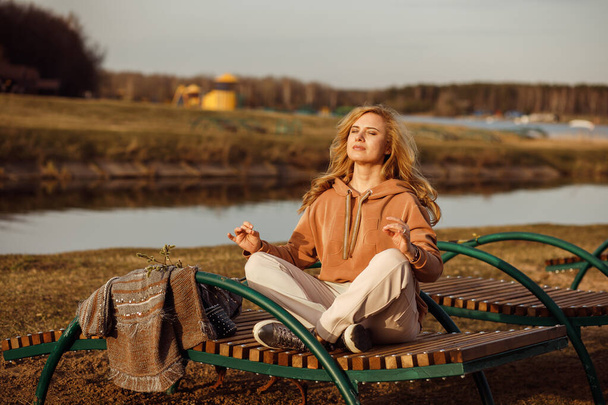 Красива блондинка сидить на лавці поруч з берегом річки в холодний сонячний день, розслабляючись, роздумуючи і насолоджуючись миром і свободою. Активність на відкритих вихідних, сільський фон природи
 - Фото, зображення