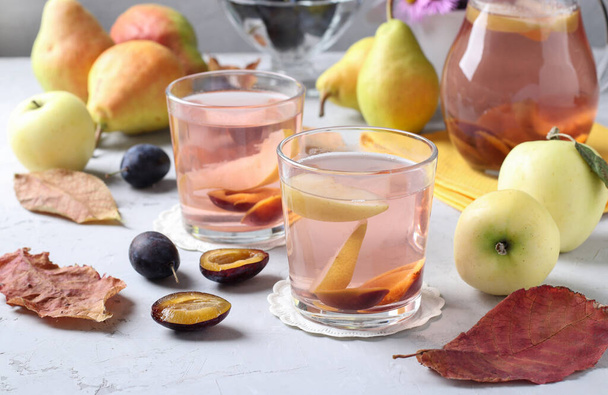Компот слив, груш и яблок в два стакана и кувшин на светло-сером фоне - Фото, изображение