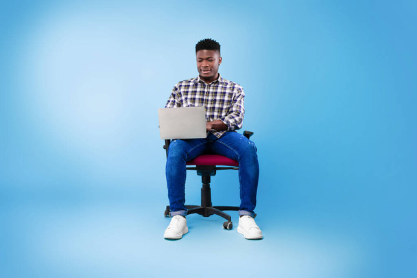 Повна довжина молодого чорного хлопця з ноутбуком, який сидить на стільці, вивчає або працює в Інтернеті на синьому фоні студії
 - Фото, зображення