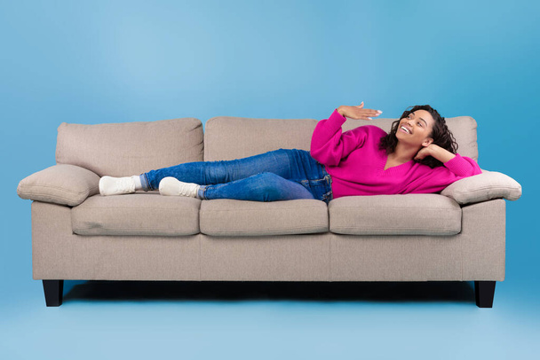 Happy young black woman lying on sofa, feeling hot, waving at herself, mockup for air conditioner or split system design - Photo, Image