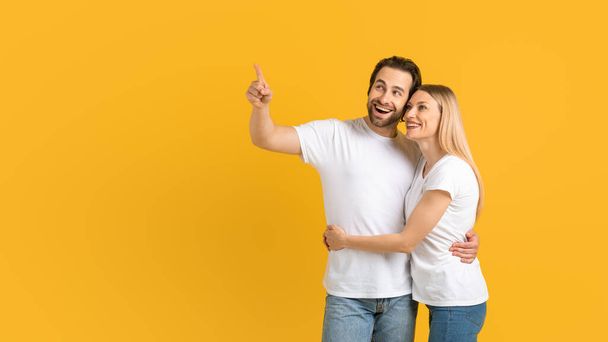 Cheerful excited young european couple in white t-shirts hugging pointing fingers at empty space - Photo, Image