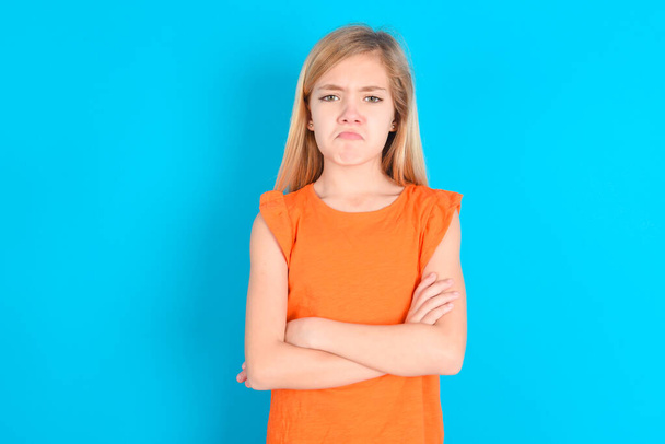 Düster unzufriedenes kleines Mädchen, das orangefarbenes T-Shirt über blauem Hintergrund trägt, schaut mit jämmerlichem Gesichtsausdruck unter der Stirn in die Kamera, macht unglückliche Fratzen - Foto, Bild