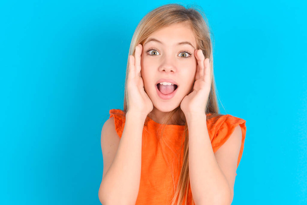 little kid girl wearing orange T-shirt over blue background Pleasant looking cheerful, Happy reaction - Photo, image