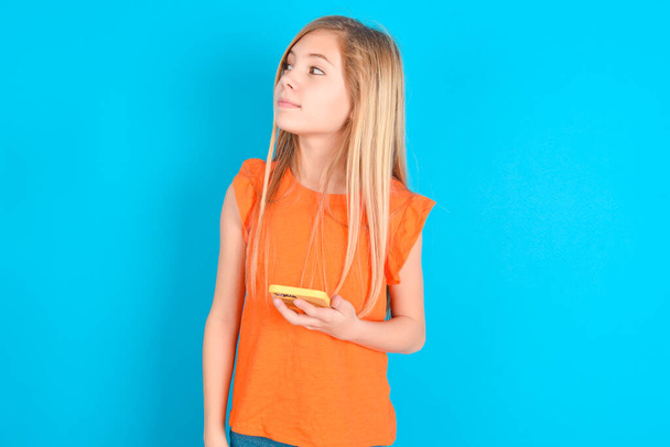niña pequeña con camiseta naranja sobre fondo azul sostiene teléfono móvil utiliza Internet de alta velocidad y las redes sociales tiene comunicación en línea. Concepto de tecnologías modernas - Foto, Imagen