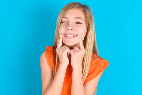 Happy little kid girl wearing orange T-shirt over blue background with toothy smile, keeps index fingers near mouth, fingers pointing and forcing cheerful smile - Valokuva, kuva