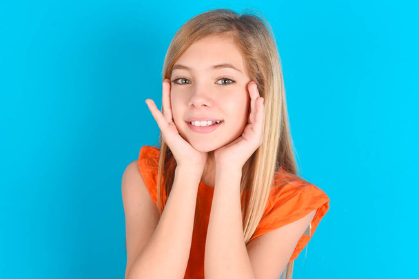 Menina criança feliz vestindo camiseta laranja sobre fundo azul toca ambas as bochechas suavemente, tem sorriso terno, mostra dentes brancos, olha positivamente diretamente para a câmera, - Foto, Imagem