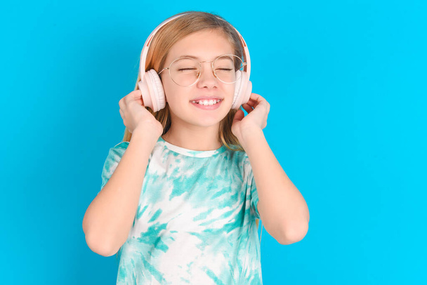 little caucasian kid girl wearing trendy T-shirt   with headphones on her head, listens to music, enjoying favorite song with closed eyes, holding hands on headset. - Photo, Image