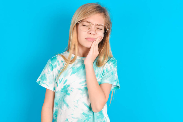 kleines kaukasisches Mädchen im trendigen T-Shirt vor blauem Hintergrund mit Zahnschmerzen im Studio - Foto, Bild