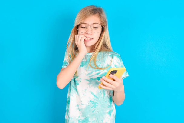 Portrait of pretty frightened little caucasian kid girl wearing trendy T-shirt over blue background chatting biting nails after reading some scary news on her smartphone. - Photo, Image