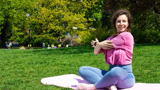Sporty beautiful young woman stretching arms during exercising outdoors before workout, sitting on mat in city park on green grass in sunny day. Preparing muscles and ligaments. Copy space for text - Photo, Image