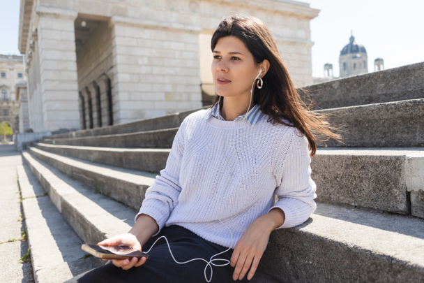 brunette woman listening music in wired earphones and holding smartphone while sitting on stairs  - 写真・画像