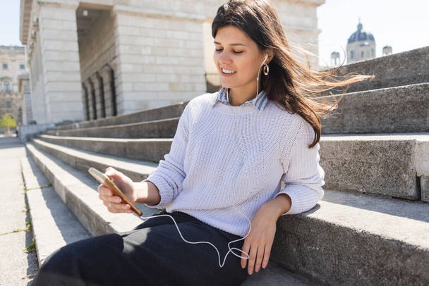 happy woman listening music in wired earphones and using smartphone while sitting on stairs  - Foto, Bild