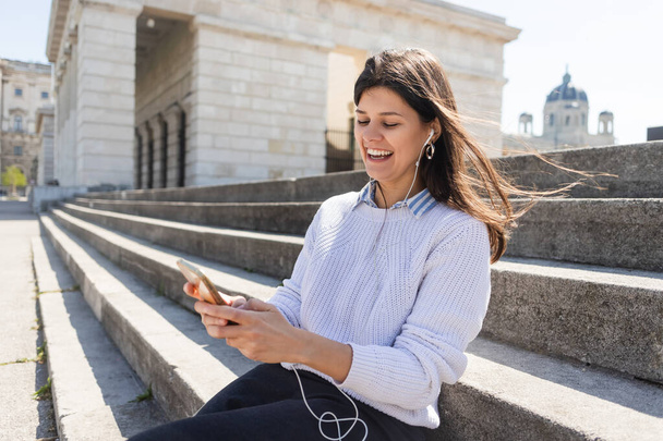 mujer alegre escuchando música en auriculares con cable y usando un teléfono inteligente mientras está sentado en las escaleras  - Foto, Imagen