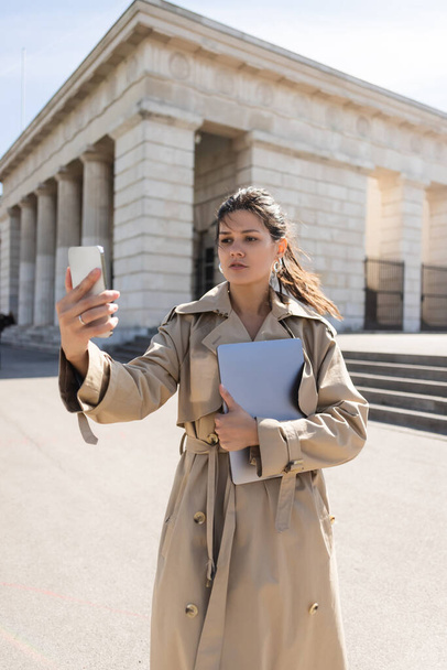 brunette woman in trench coat holding laptop and looking at smartphone outside - 写真・画像