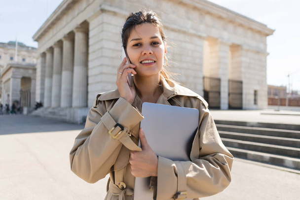 pleased freelancer in trench coat holding laptop and talking on smartphone outside - Photo, image