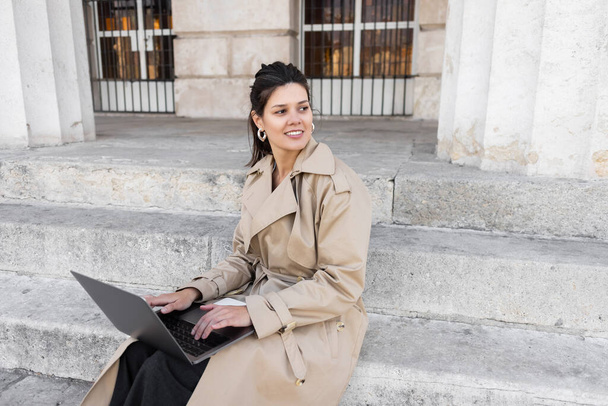 pleased woman in trench coat sitting on concrete stairs and working remotely on laptop  - Photo, Image