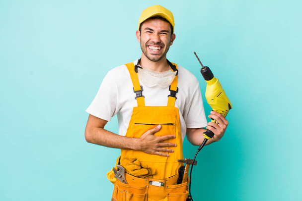 young adult hispanic man laughing out loud at some hilarious joke. worker and drill concept - Foto, afbeelding