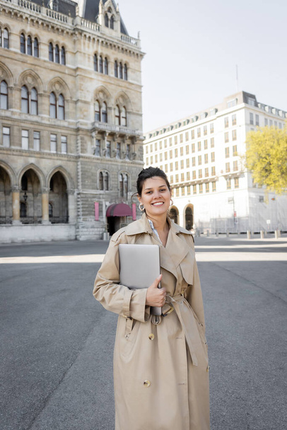 happy freelancer in beige trench coat walking with laptop on street of vienna  - Foto, Imagen