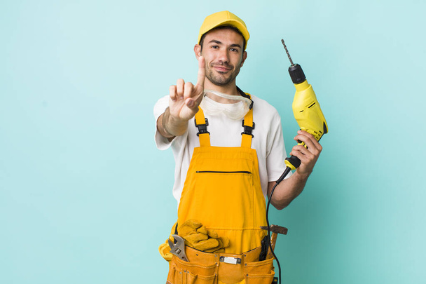 young adult hispanic man smiling and looking friendly, showing number one. worker and drill concept - Фото, зображення