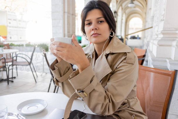 mulher morena em casaco de trincheira ouvindo música e segurando copo perto de smartphone e laptop no terraço do café - Foto, Imagem