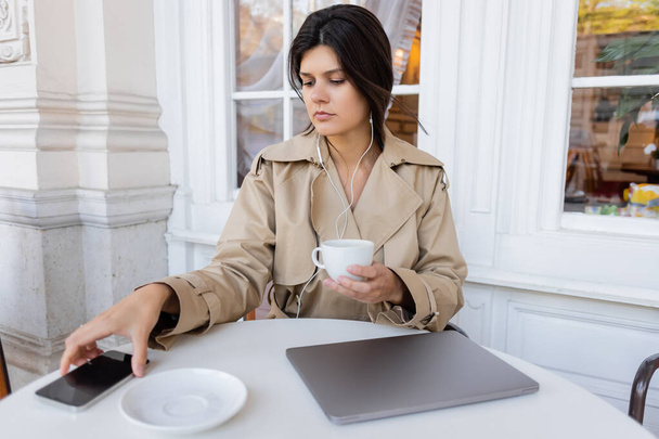 brunette woman in trench coat listening music and holding cup while reaching smartphone on table in cafe terrace - Photo, Image
