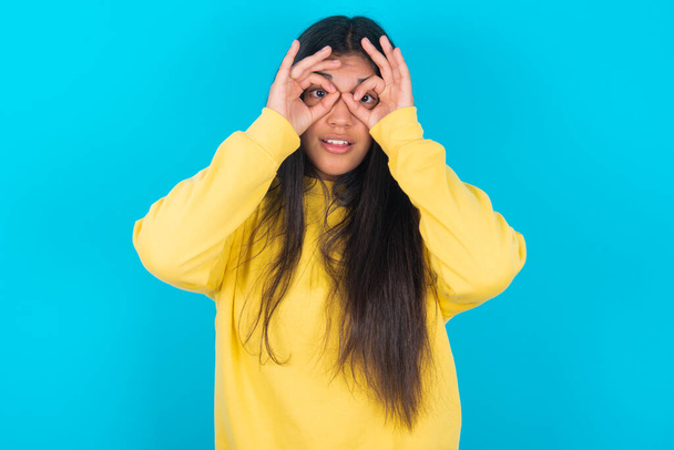 игривая возбужденная латиноамериканка в жёлтой толстовке на синем фоне показывает знак ОК с обеими руками на глазах, притворяясь, что носит очки. - Фото, изображение