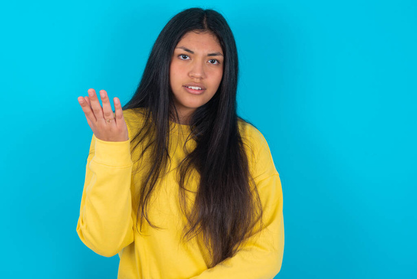 Studio shot of frustrated  latin woman wearing yellow sweatshirt over blue background gesturing with raised palm, frowning, being displeased and confused with dumb question. - 写真・画像
