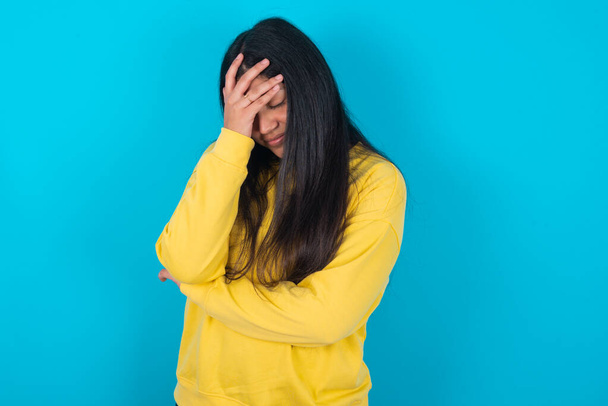 latin woman wearing yellow sweatshirt over blue background making facepalm gesture while smiling amazed with stupid situation. - Zdjęcie, obraz