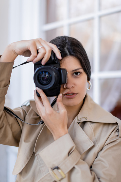 brunette photographer in trench coat taking photo on digital camera  - Foto, afbeelding