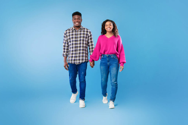 Volledige lengte van de millennial Afro-Amerikaanse paar lopen, hand in hand, glimlachen op camera op blauwe studio achtergrond - Foto, afbeelding