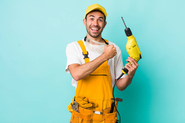 young adult hispanic man feeling happy and facing a challenge or celebrating. worker and drill concept - Photo, Image