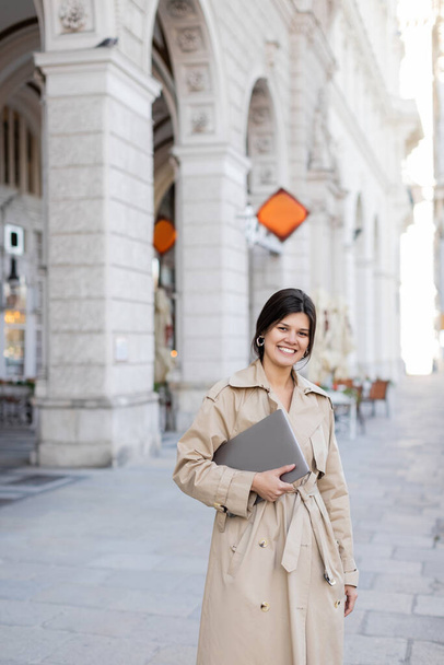 happy woman in trench coat walking with laptop on street of vienna  - Foto, Bild