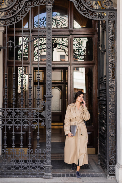 žena v trenč kabát mluví na smartphone a stojí s notebookem v blízkosti padělaných dveří - Fotografie, Obrázek