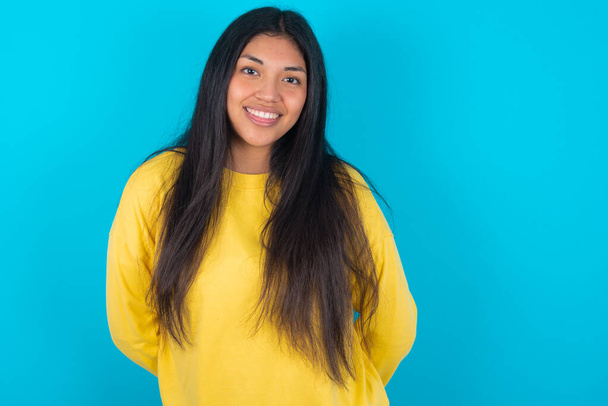 femme latine portant un sweat-shirt jaune sur fond bleu avec un visage heureux et drôle souriant et montrant la langue. - Photo, image