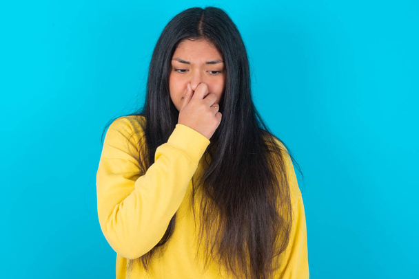 mujer latina con sudadera amarilla sobre fondo azul sosteniendo su nariz debido a un mal olor. - Foto, Imagen