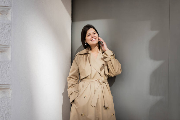 happy woman in trench coat standing with hand in pocket and talking on smartphone  - 写真・画像