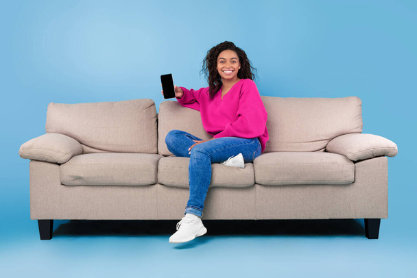 Jeune femme noire positive assise sur le canapé avec téléphone portable sur fond bleu studio, maquette pour application ou site Web - Photo, image