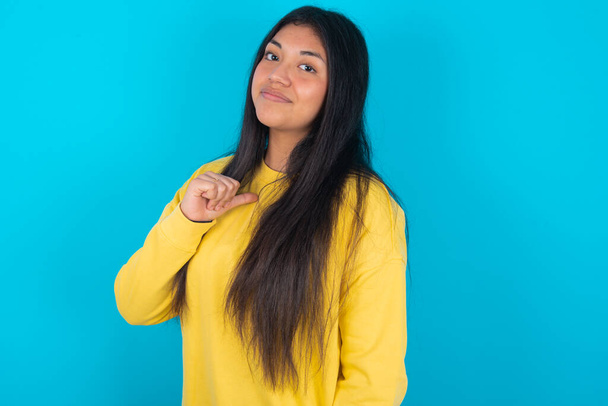 Closeup of cheerful latin woman wearing yellow sweatshirt over blue background looks joyful, satisfied and confident, points at himself with thumb. - Photo, Image