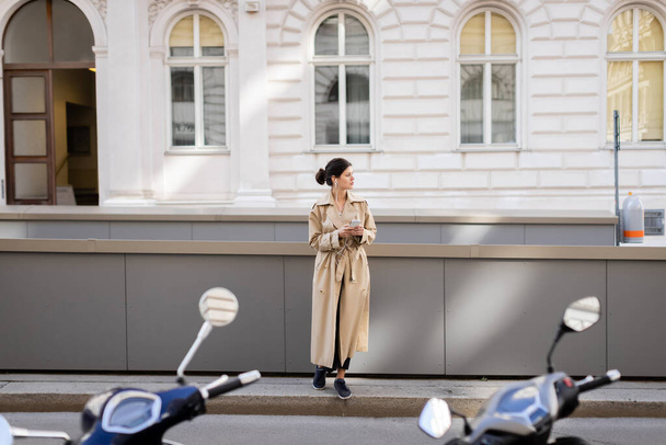 full length of brunette woman in wired earphones holding mobile phone and listening music on street in vienna - 写真・画像